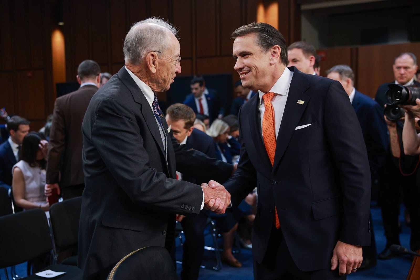 Senator Chuck Grassley shaking hands with Todd Blanche in a Senate hearing room, with photographers behind them.
