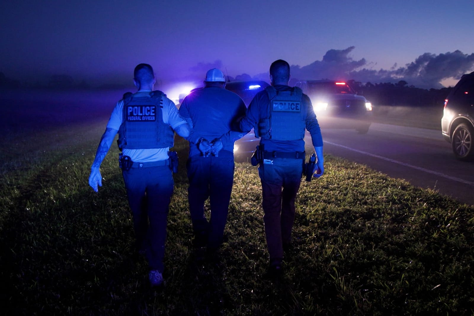 Two ICE officers walk a handcuffed man away from the camera at dusk beside a highway, police lights behind them.