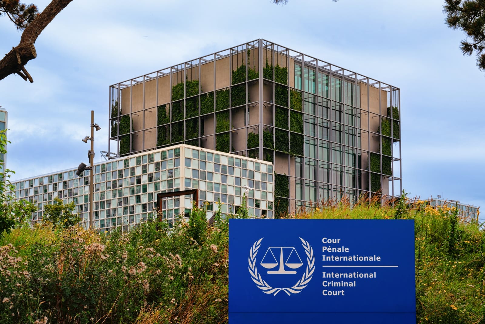 The International Criminal Court’s glass-and-green building in The Hague, behind a blue sign reading International Criminal Court.