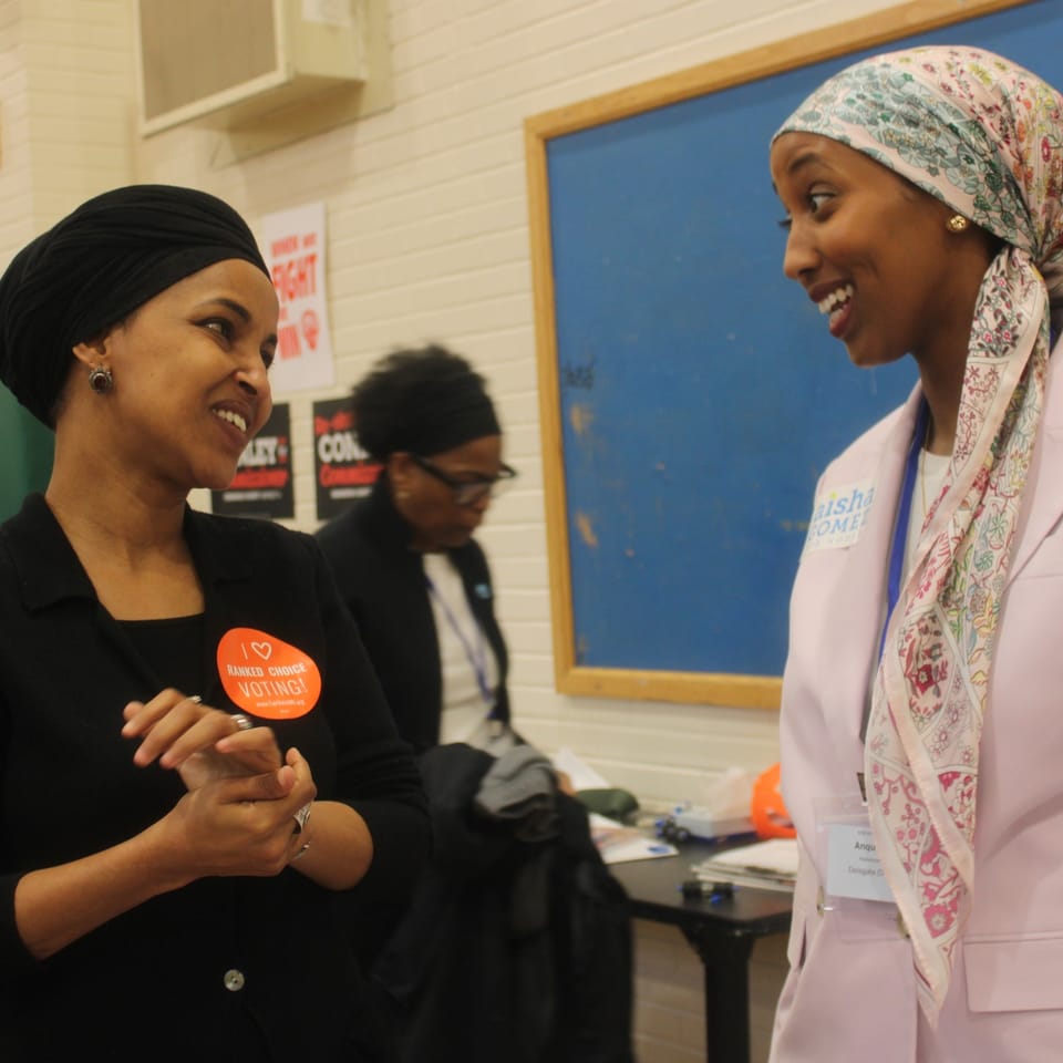 Ilhan Omar speaking into a microphone at a lectern, with campaign signs on the wall behind her.