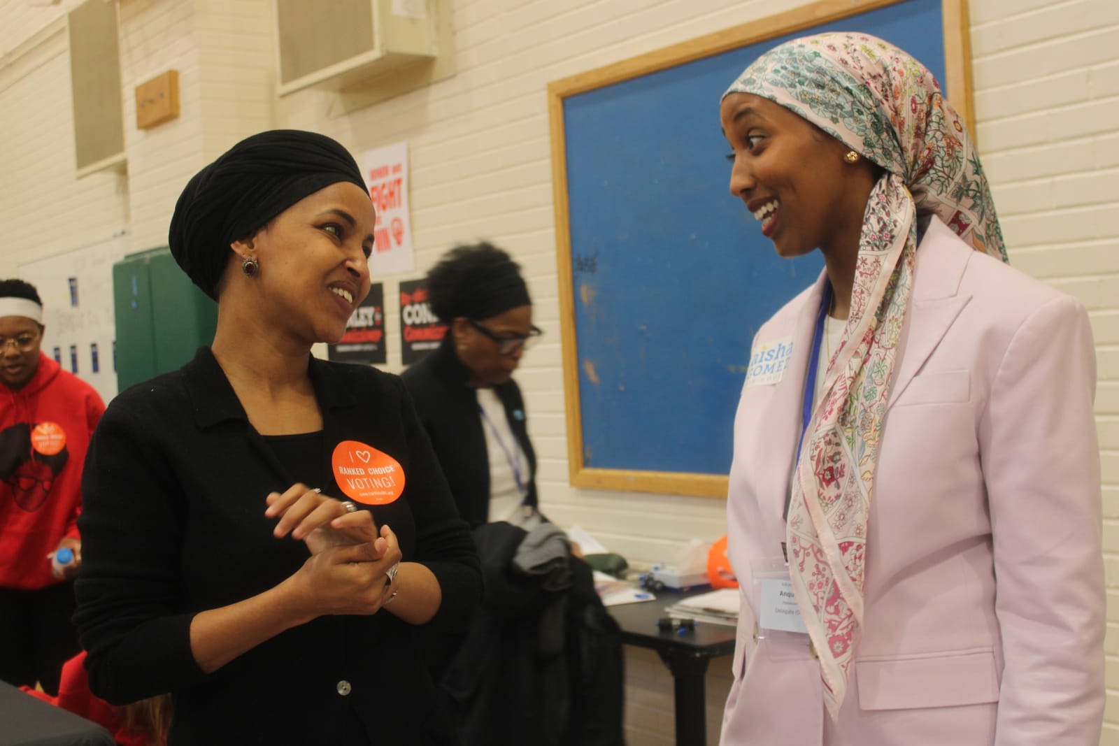 Ilhan Omar speaking into a microphone at a lectern, with campaign signs on the wall behind her.